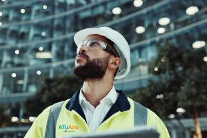 Persona equipada con casco de seguridad, gafas de protección y chaleco reflectante realizando labores de supervisión técnica frente a un edificio moderno. En la prenda aparece el logotipo de ATuAire by Iberdrola.