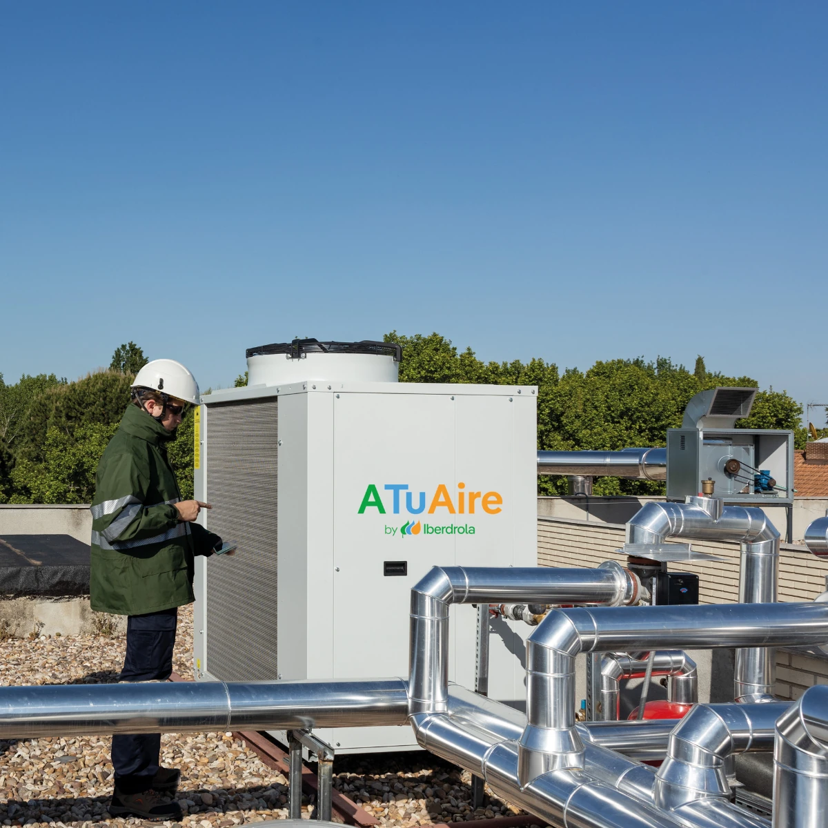 Técnico inspeccionando una unidad de aerotermia o bomba de calor ATuAire by Iberdrola instalada en una cubierta técnica, rodeada de tuberías metálicas aisladas y equipos auxiliares de climatización.