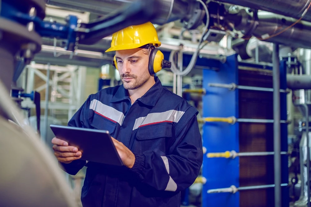 Hombre en traje de trabajo con casco mirando una tablet en una sala de máquinas.