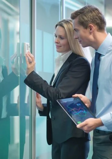 Dos profesionales revisan la configuración de un termostato o sistema de control de climatización instalado en un edificio, mientras consultan información en una tableta digital.