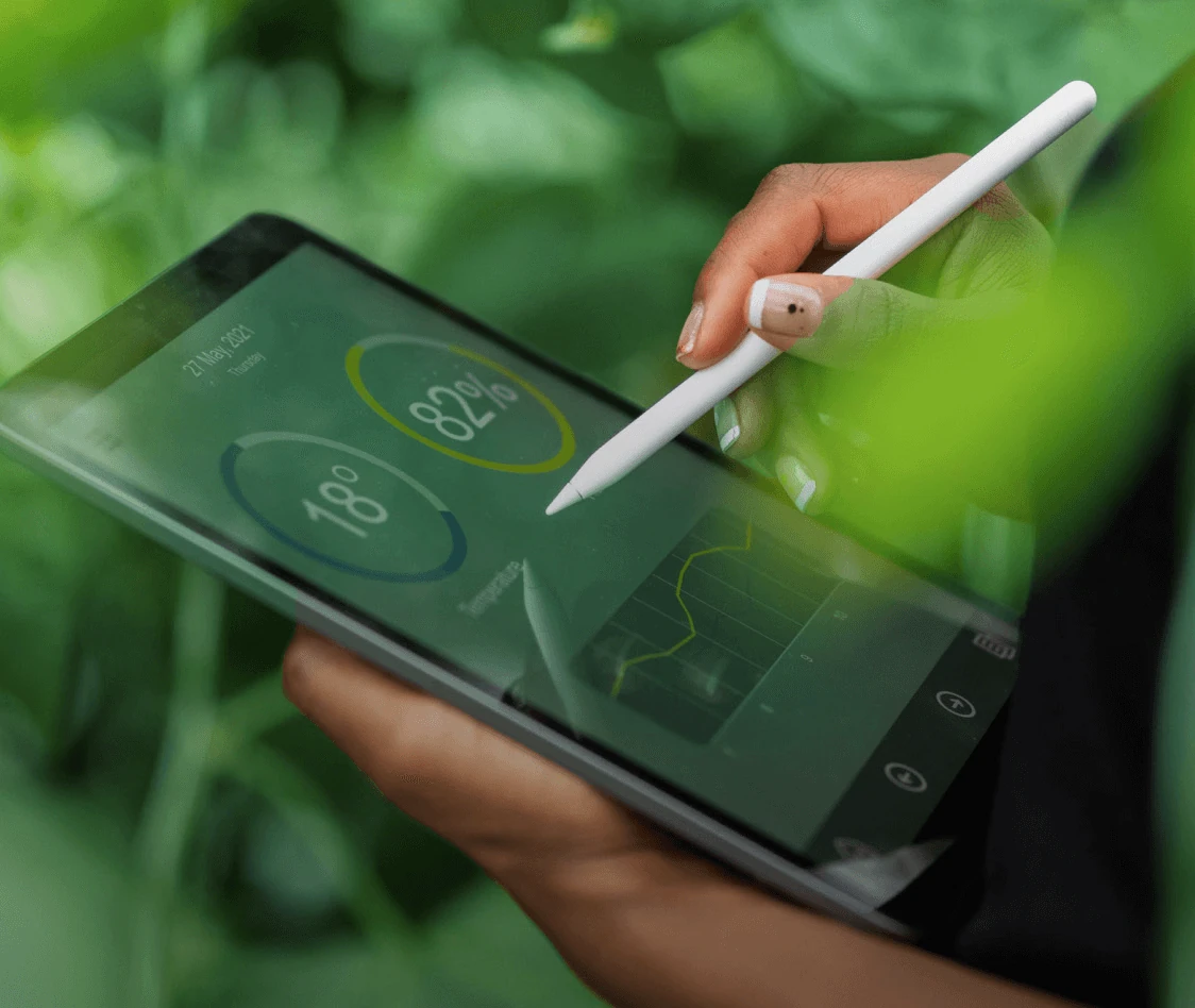 Persona utilizando un lápiz digital sobre una tableta que muestra gráficos e indicadores de rendimiento, rodeada de vegetación.