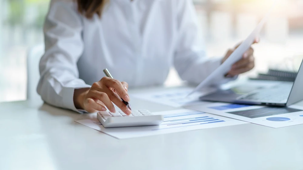 Persona sentada en un escritorio utilizando una calculadora mientras revisa documentos con gráficos financieros; junto a los papeles hay un ordenador portátil abierto.