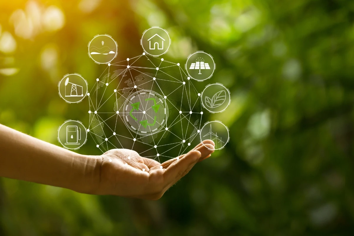 Mano abierta sosteniendo una representación digital del planeta Tierra rodeada de iconos relacionados con sostenibilidad, reciclaje, energías renovables y eficiencia energética, sobre un fondo natural desenfocado en tonos verdes.
