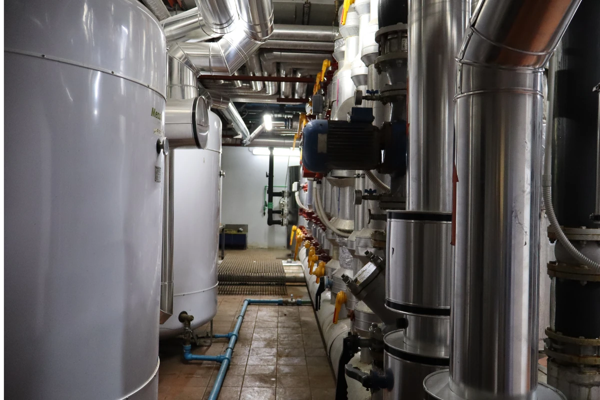 Sala técnica con una instalación de climatización o calefacción compuesta por tuberías metálicas aisladas, válvulas, intercambiadores de calor, bombas y depósitos industriales conectados mediante una red de conductos.