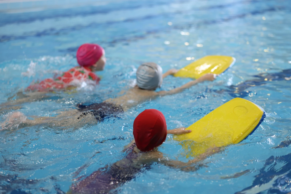 Grupo de niños realizando una actividad de aprendizaje de natación en una piscina cubierta, utilizando tablas flotantes amarillas para practicar el desplazamiento en el agua.