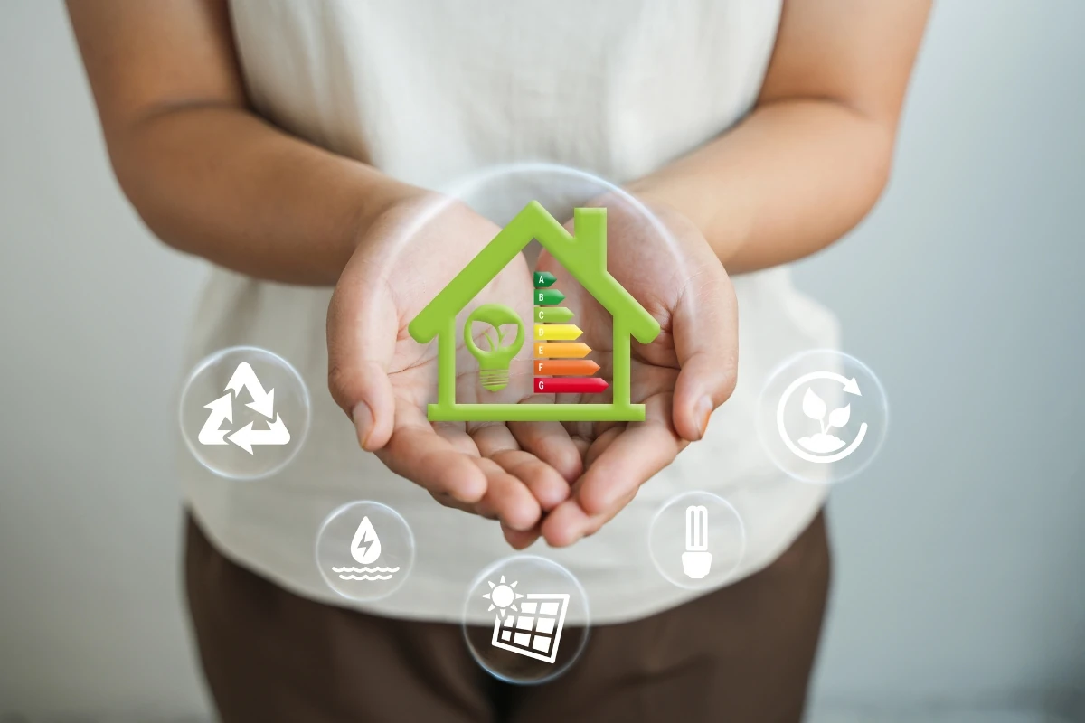 Manos sosteniendo una representación de una vivienda eficiente energéticamente, con una etiqueta de clasificación energética de la A a la G y una bombilla ecológica en su interior. Alrededor aparecen iconos relacionados con reciclaje, energías renovables, ahorro energético e iluminación eficiente.