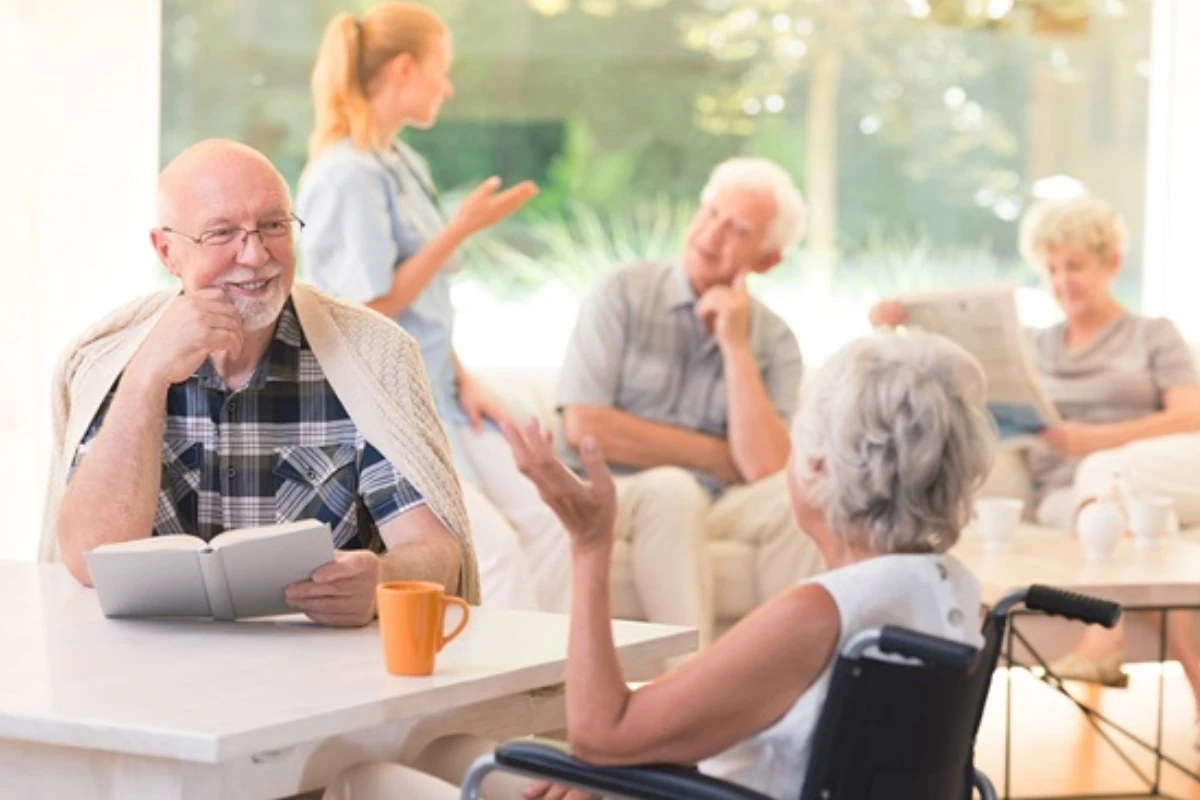 Sala común de un centro residencial o de atención a personas mayores, donde varias personas participan en actividades sociales y de lectura. En primer plano, una persona lee un libro mientras conversa con otra persona usuaria de silla de ruedas.