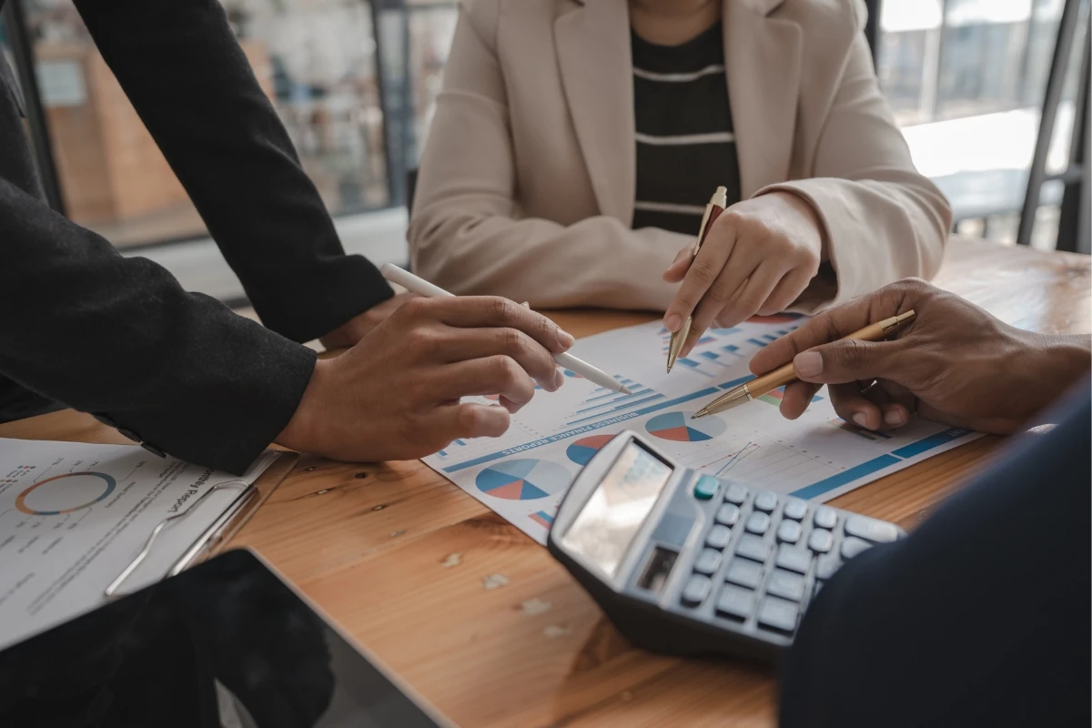 Varias personas analizan documentos con gráficos y datos financieros durante una reunión de trabajo. Sobre la mesa hay informes impresos, una calculadora, una tableta electrónica y varios bolígrafos utilizados para revisar los resultados.