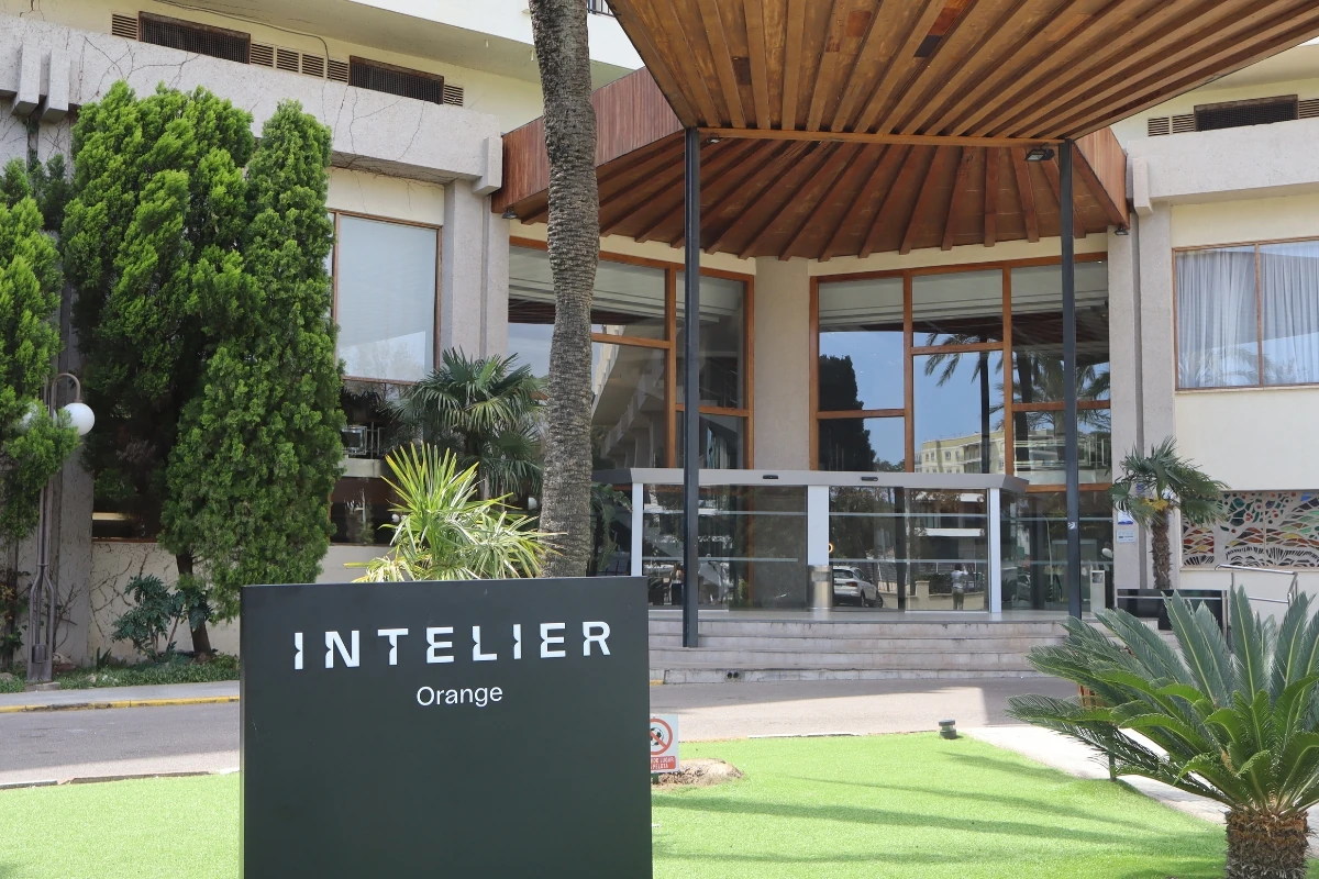 Entrada principal del hotel Intelier Orange, con un acceso cubierto por una estructura de madera, puertas automáticas acristaladas y zonas ajardinadas con palmeras y vegetación ornamental.