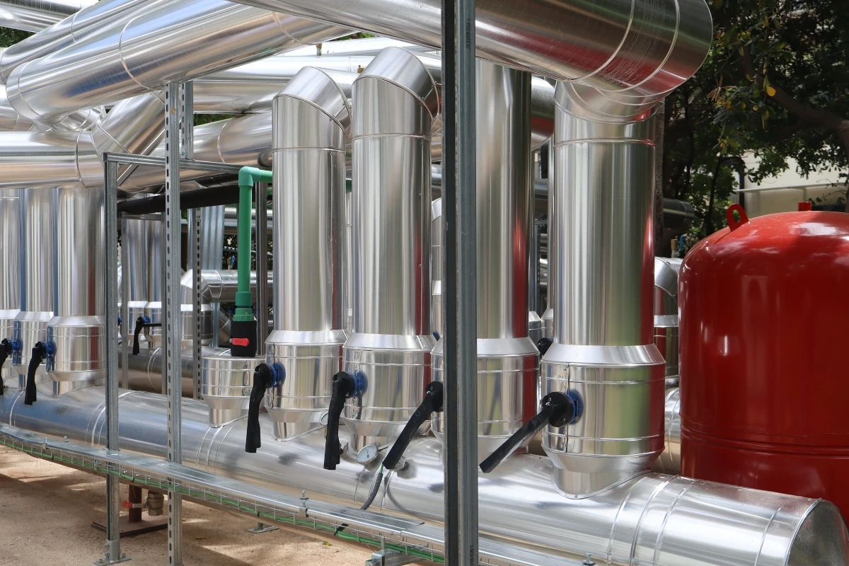 Red de tuberías industriales aisladas térmicamente en una instalación de climatización o producción de energía, formada por conductos metálicos de gran diámetro, válvulas de control y un depósito de expansión de color rojo.