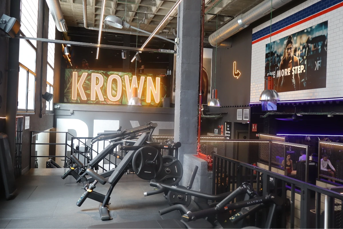 Zona de entrenamiento de Krown Fitness equipada con máquinas de musculación, espacios diáfanos y elementos de diseño industrial. Al fondo destaca la señal luminosa con el nombre del gimnasio.