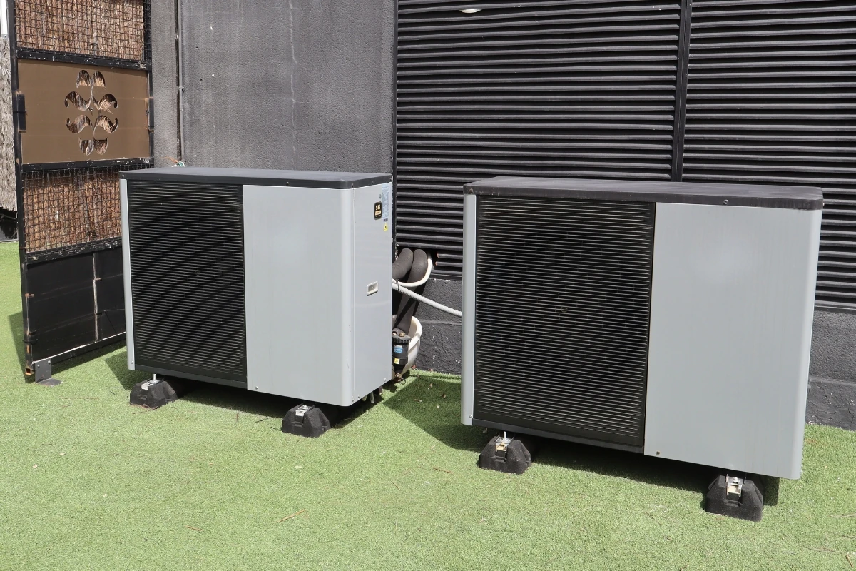 Dos unidades exteriores de bomba de calor o climatización instaladas en el exterior de un edificio, conectadas mediante tuberías y situadas sobre soportes antivibración junto a una rejilla de ventilación.