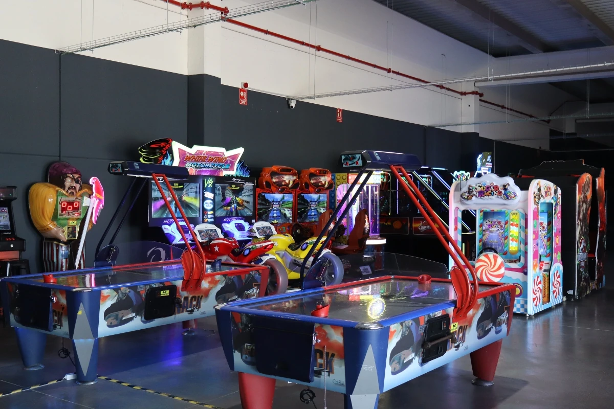 Zona de juegos recreativos en el interior de un centro de ocio, equipada con mesas de air hockey, motocicletas de simulación y diversas máquinas arcade iluminadas con luces de colores.