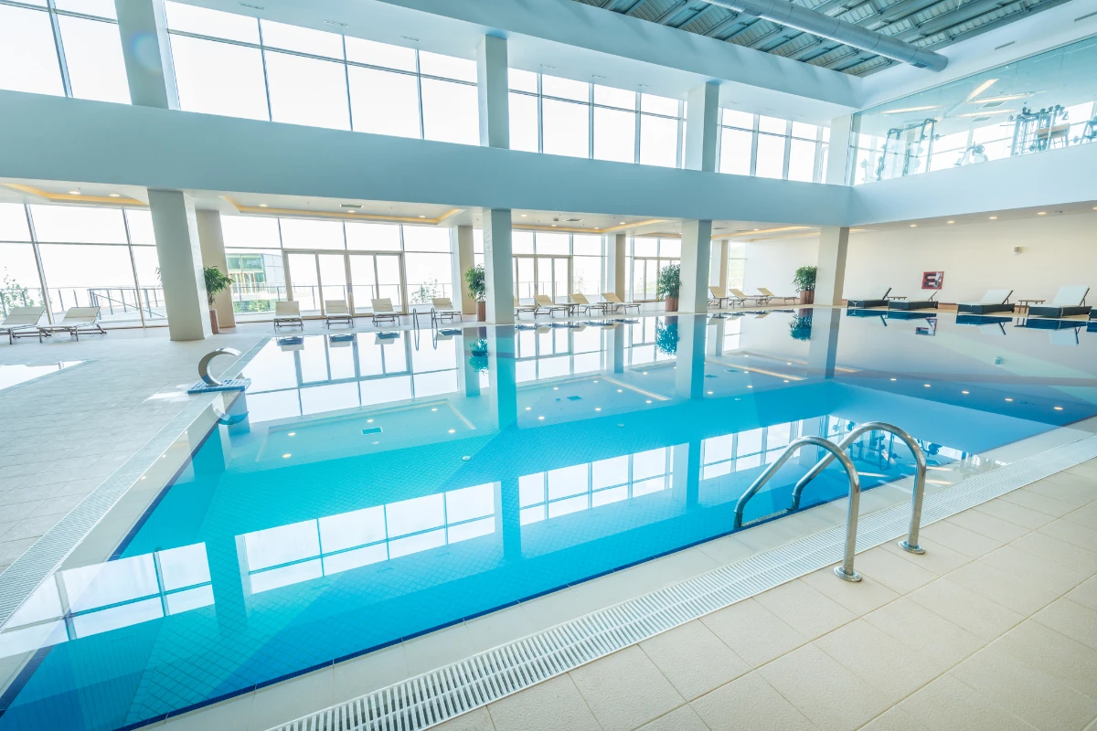 Piscina climatizada interior de un centro de bienestar o complejo hotelero, con amplios ventanales, zona de descanso con tumbonas y abundante luz natural reflejada en el agua.