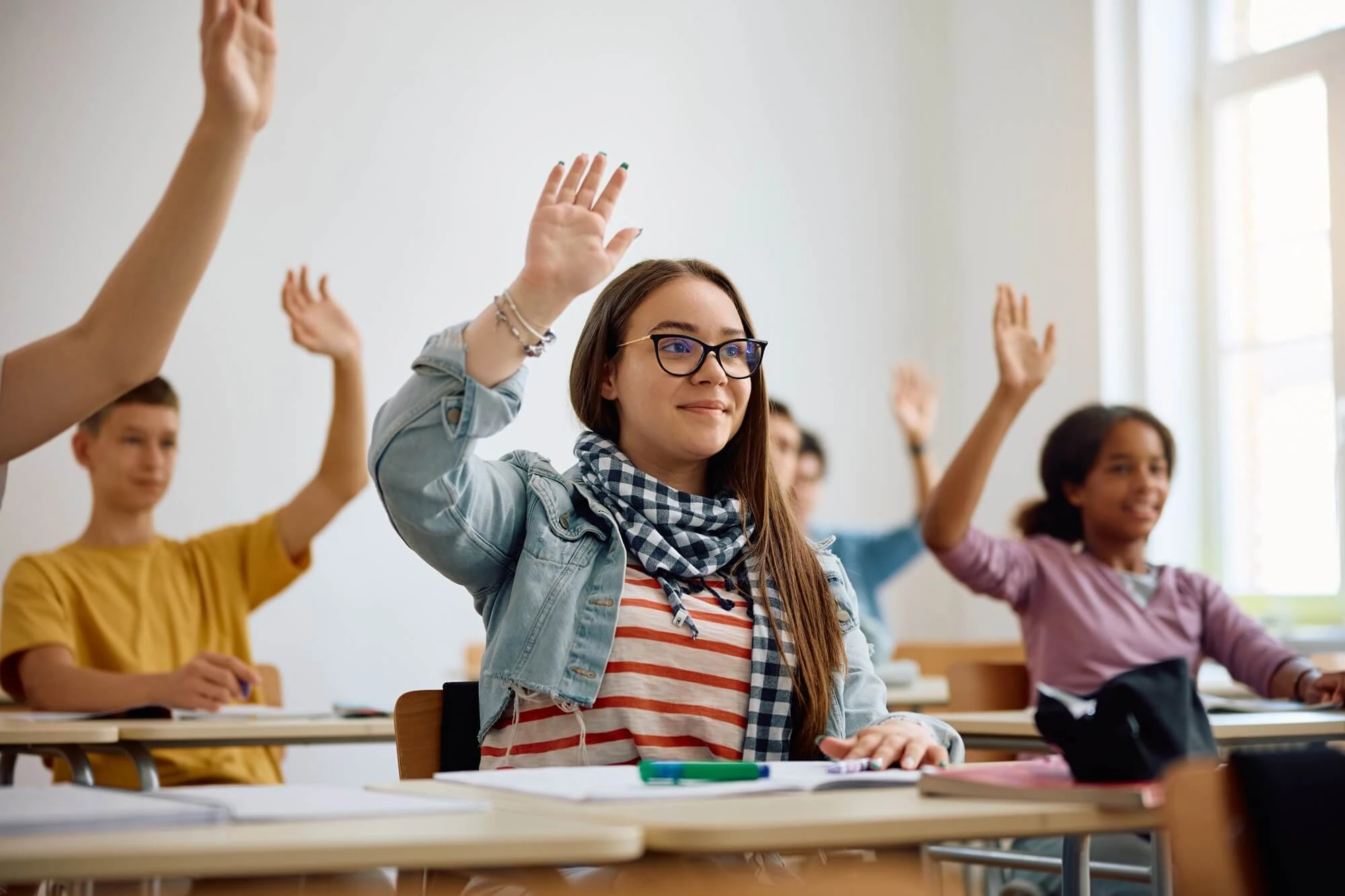 Aula educativa donde varios estudiantes levantan la mano para participar en una actividad o responder a una pregunta. En primer plano, una estudiante con gafas está sentada en su mesa con material escolar mientras participa en la clase.