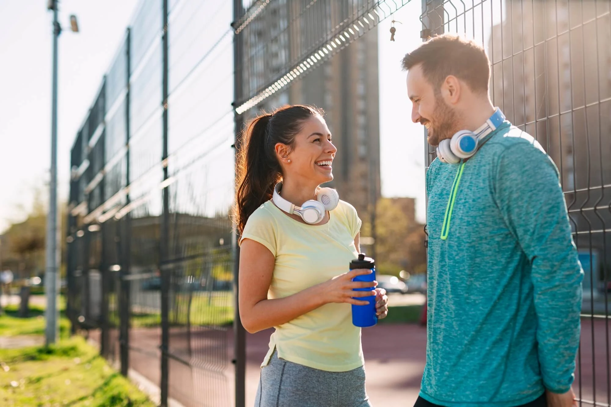 Dos personas con ropa deportiva conversan junto a una pista o instalación deportiva exterior, con auriculares alrededor del cuello y una botella reutilizable en primer plano.