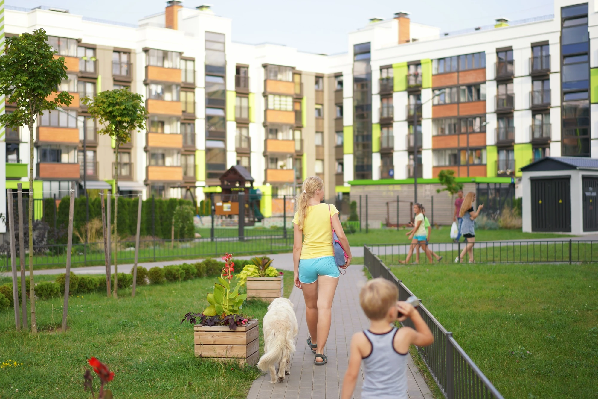 Conjunto residencial de viviendas multifamiliares con zonas ajardinadas, áreas peatonales y espacios comunes. En primer plano, varias personas pasean por los jardines y una persona camina junto a un perro por una senda interior del complejo.