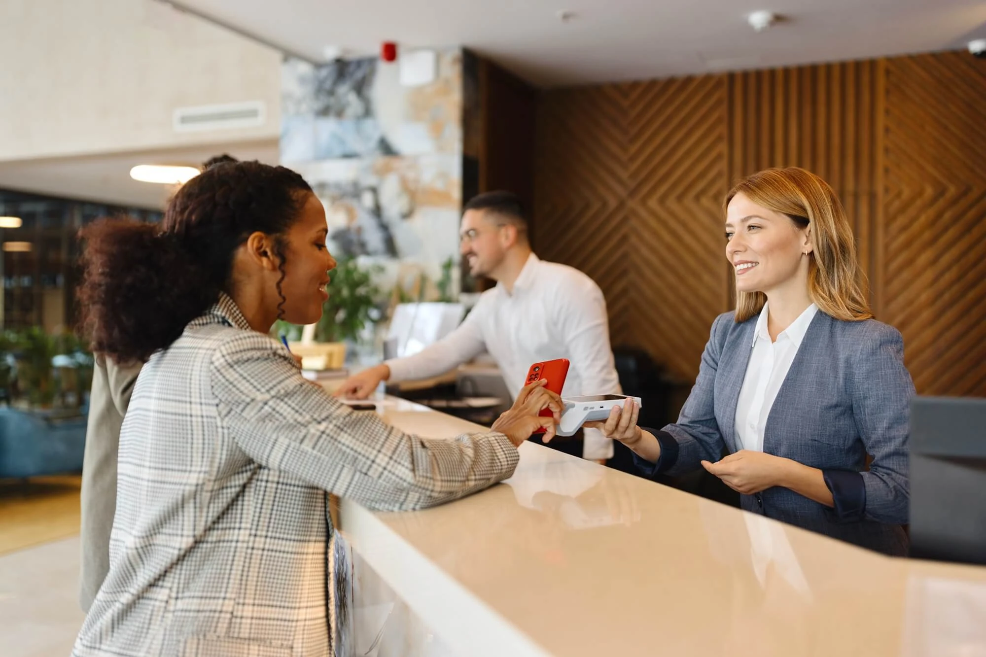 Recepción de un hotel donde una persona realiza un pago o registro utilizando un dispositivo móvil, mientras el personal de atención al cliente opera un terminal de cobro en el mostrador.
